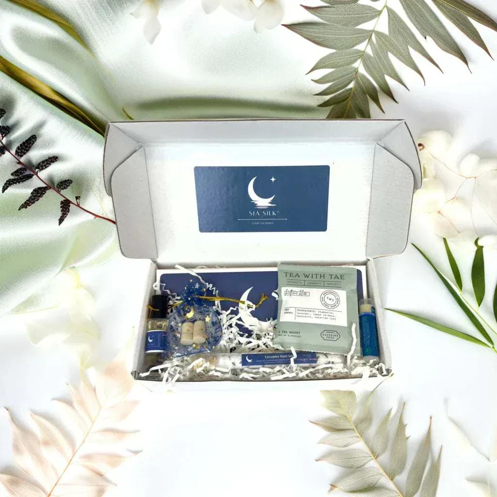Open gift box containing Sia Silk® Sleep Kit with silk sleep mask, lavender essential oil roller, lip balm, bath salts, bedtime tea, and foam ear plugs in eco-friendly packaging.
